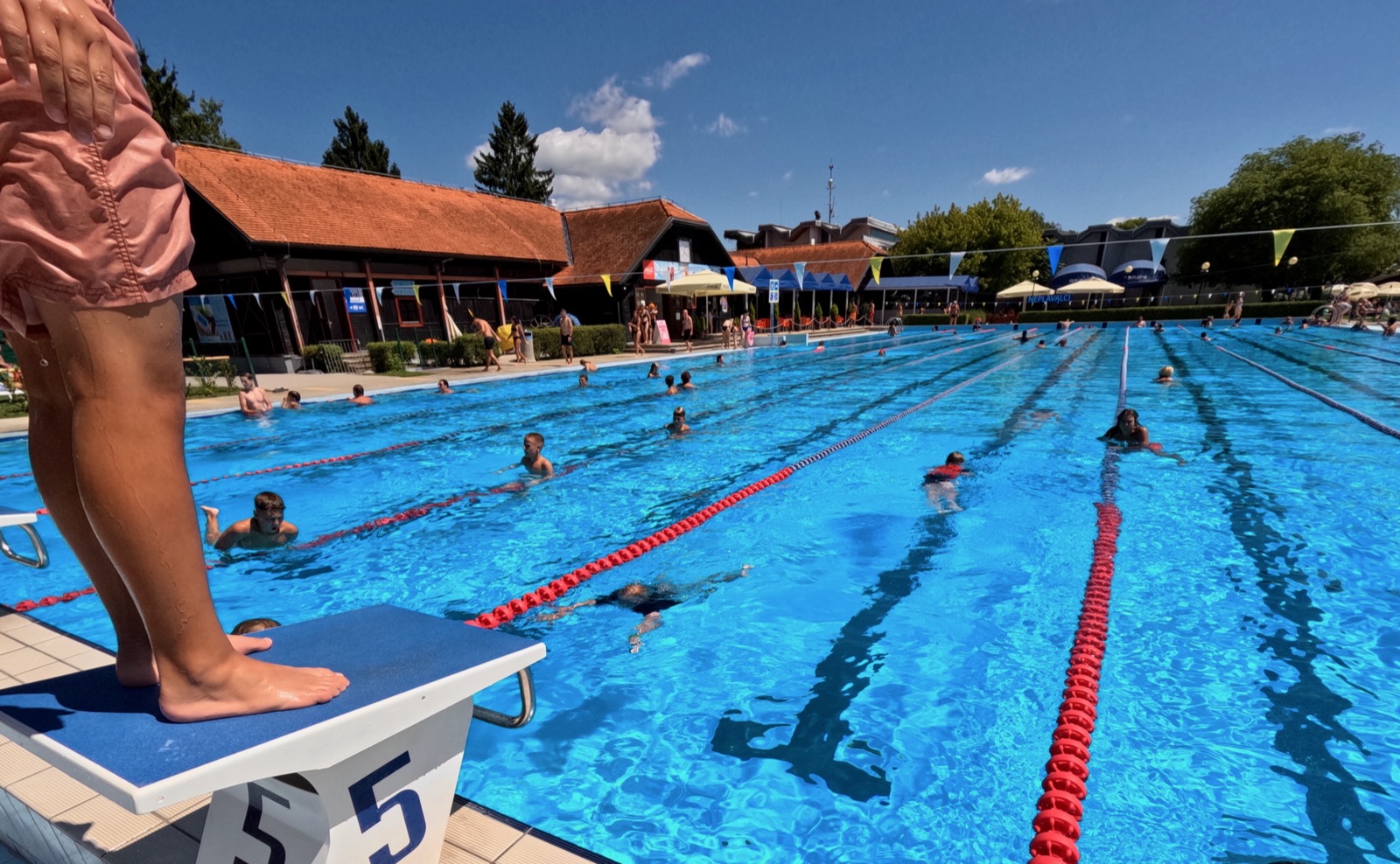 Person standing on diving board above swimming pool with people swimming.